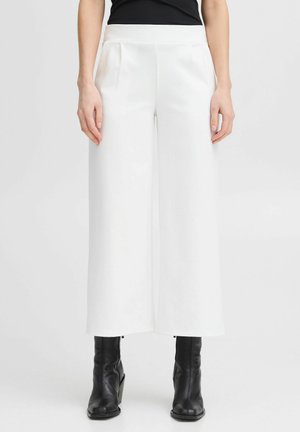 Pantalons larges blancs avec une texture lisse, dotés d'une taille haute et d'un devant plissé. Associés à des bottines à talons noirs.