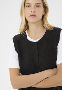 Gilet lavorato a maglia nero con scollo rotondo, chiusura con bottoni e tessuto testurizzato, indossato sopra una camicia bianca a maniche corte.