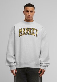Szürke pulóver lekerekített nyakrésszel és oversized fazonnal. "MARKET" felirat kamuflázs mintával a mellkason. Rugalmas mandzsetták és alj.