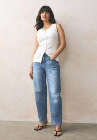 Weißes, ärmelloses, geripptes Oberteil mit Knöpfen, kombiniert mit hellblauen, weit geschnittenen Jeans und schwarzen Sandalen mit Riemchen. Neutrale Hintergrundstruktur.