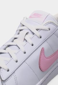 Baskets blanches Nike avec logo Swoosh rose et marquage rose sur la languette, lacets blancs noués en motif croisé standard.