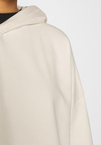 Sudadera con capucha beige con un gran bolsillo frontal, que presenta una textura suave y detalles de costuras visibles a lo largo de las costuras y la capucha.