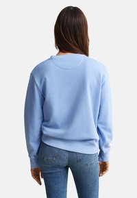 Άτομο με μακριά σκούρα μαλλιά που κοιτάει από την πλάτη, φοράει ανοιχτό μπλε φούτερ και μπλε τζιν μπροστά σε απλό ανοιχτό φόντο.