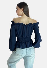 Marineblaue Off-Shoulder-Bluse mit gerüschtem Halsausschnitt und langen Ärmeln; verfügt über ein gesmoktes Taillendetail und eine fließende Silhouette. Kombiniert mit hellen Jeans.