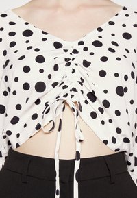 Top cropped branco com bolinhas pretas, apresenta um decote em V profundo, centro franzido com laços e mangas curtas. Combinado com calças pretas de cintura alta.