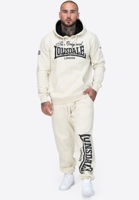 Sudadera cremosa con el texto negro "The Original Lonsdale London", combinada con unos joggers crema a juego que presentan un logotipo en la pierna izquierda. Zapatillas blancas.