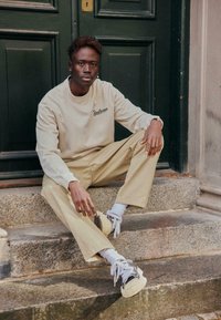 Sweat-shirt beige avec broderie "Soulscape", associé à un pantalon beige clair. Baskets grises et blanches avec lacets visibles, assis sur des marches en pierre.