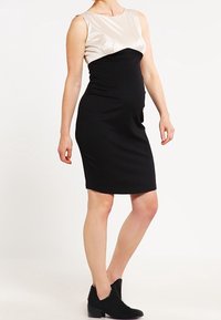 Robe noire longueur genou avec une silhouette ajustée, présentant un haut en satin beige clair et une jupe en tissu noir lisse. Portée avec des bottines.