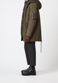 Man som bär en olivgrön parkas med huva, mörkgrå cropped byxor och svarta snörskor, stående i profil mot vit bakgrund.