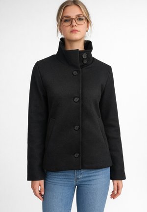 Femme portant un manteau noir boutonné à col montant et un jean bleu, debout devant un fond blanc.