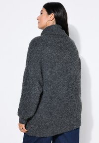 Grauer, flauschiger Strickpullover mit hohem Kragen, lockerer Passform und langen Ärmeln, der eine strukturierte Oberfläche und einen längeren hinteren Saum aufweist.