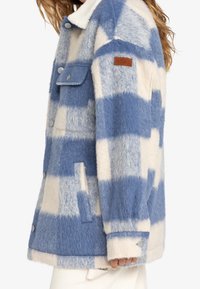 Frau trägt eine blau-weiße kariert Fleecejacke mit Metallknöpfen und einem braunen "Roxy"-Patch auf dem oberen Arm.