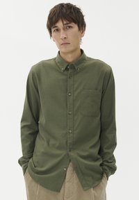 Olivno zelena srajca z gumbi, z žepom, dolgimi rokavi in ovratnikom. Narejena iz teksturirane tkanine z vidnimi šivi in gumbi.