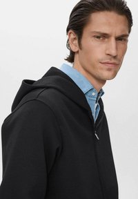 Sudadera negra con cremallera y una textura suave, que cuenta con una capucha ajustable. Llevada sobre una camisa de botones azul claro, tiene un diseño moderno y ajustado.