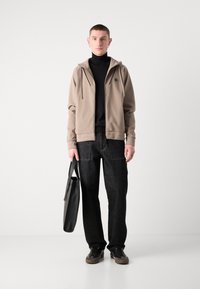Béžová mikina na zips s logom, čierny rolák, tmavé džínsy a šedé tenisky. Drží čiernu tašku. Jednoduchý, neformálny outfit.