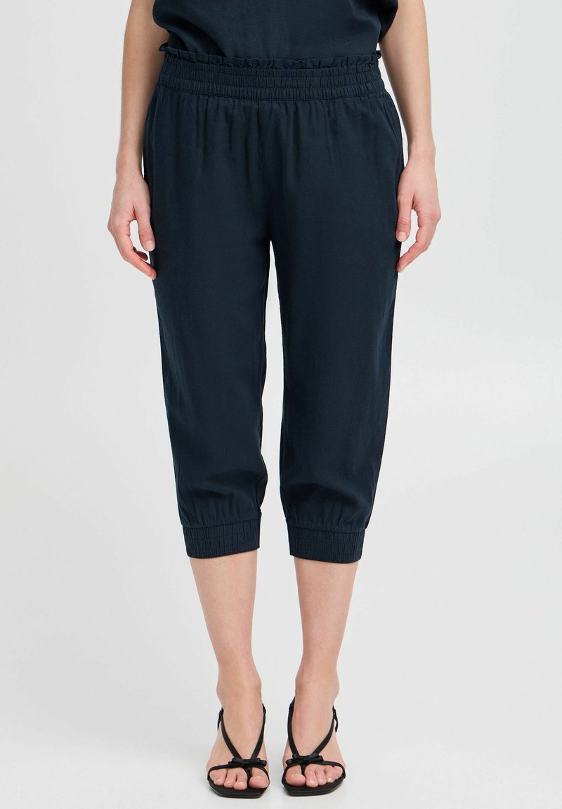 Femme portant un pantalon court noir avec taille et poignets froncés élastiqués, associé à des sandales noires à talons, debout devant un fond uni.