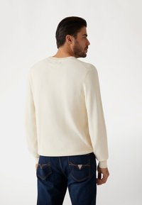 Un pull en crème clair avec des manches longues et des poignets côtelés, associé à un jean bleu foncé avec des coutures contrastées et un détail de logo.