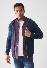 Marineblå zip-up hættetrøje med en kengurulomme, båret over en rød og marineblå ternet skjorte og en hvid t-shirt. Modellen poserer med hånden hævet.