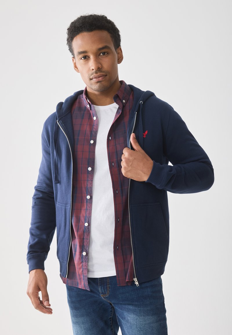 Navy zip-up hoodie with a kangaroo pocket, worn over a red and navy checkered shirt and white t-shirt. Model is posing with hand raised.
