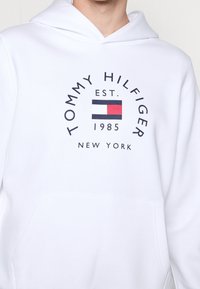 Sudadera blanca con bolsillo frontal, capucha ajustable y un logo impreso con un diseño de bloques de color que incluye acentos en rojo, azul y blanco.