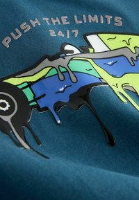 Spalvingas stilizuotas lenktyninis automobilis su dažų lašais ant mėlyno audinio, lydimas užrašo "KELKITE RIBAS 2017."