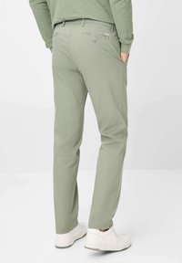 Light green chinos with a straight cut, featuring back pockets and a button detail, paired with white sneakers on a plain background.