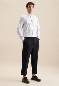 Helles blaues und weißes kariertes Hemd, zugeknöpft, mit einem Kragen. Getragen mit gefalteten schwarzen Hosen und schwarzen Dressschuhen. Einfarbiger Hintergrund.