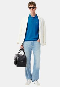 Blazer bianco, polo blu, jeans chiari, borsa a tracolla nera con dettagli bianchi, sneakers bianche e occhiali da sole neri.
