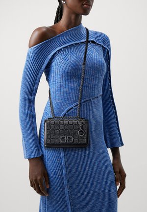 Vrouw draagt een blauwe geribbelde off-shoulder jurk met knoopdetails en een zwarte crossbodytas met patroon en kettingriem.