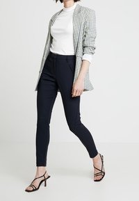 Rutig kavaj i vitt och grönt, vit polotröja, marinblå slim-fit byxor och svarta klackskor med remmar. Kavajen har en avslappnad passform och rullade ärmar.