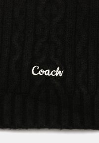 Sort strikket tørklæde med et tekstureret kabelmønster, med "Coach" broderet i hvid tråd nederst til højre.
