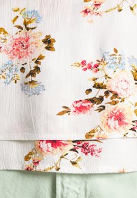 Tecido com padrão floral, apresentando flores rosa, azuis e douradas sobre um fundo branco, sobreposto a uma textura verde menta. Aparência amarrotada.