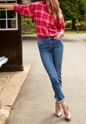 Vrouw die buiten staat met één hand in haar zak, draagt een roze geruite blouse, opgerolde blauwe jeans en nudekleurige pumps met een spits model.