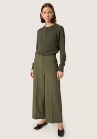 Olivgrüner gerippter Cardigan mit vorderen Knöpfen, kombiniert mit olivegrünen weiten Hosen. Das Model trägt dunkelbraune Ballerinas.