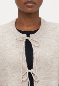 Beige knitted cardigan with a rounded neckline, featuring a tie closure at the front and ribbed detailing along the edges.
