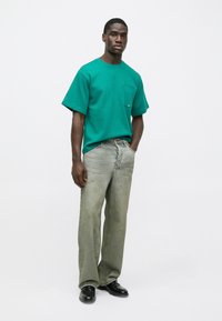 Πράσινη κοντομάνικη μπλούζα με τσέπη στο στήθος, σε συνδυασμό με ανοιχτό γκρι παντελόνι φαρδιάς γραμμής και μαύρα παπούτσια. Λείος υφασμάτινος ιστός και casual εφαρμογή.