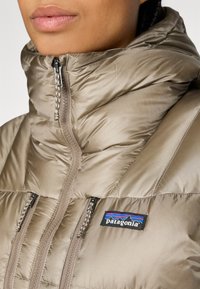 Dik beige jack met hoge kraag, ritssluiting aan de voorkant en twee ritszakken. Bevat een Patagonia-logo op het linkerborstgedeelte.