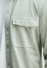 Lichtgroen shirt van zacht stof, voorzien van een borstzak met flap en een logo, met knopen en een lichte textuur zichtbaar.
