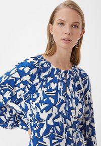 Blaue und weiße Bluse mit abstraktem Muster, langen Ärmeln, gerafftem Ausschnitt und einem Schlitz vorne. Glatte Stoffoberfläche.