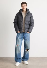 Pufferjack in donkergrijs met een ritssluiting, capuchon en doorgestikt ontwerp. Gecombineerd met versleten lichtblauwe jeans en witte sneakers.