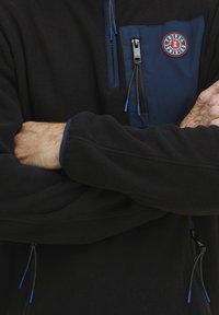 Juodas flisinis megztinis su užtrauktuko krūtine kišenė, mėlynos spalvos, su mėlynomis užtrauktuko traukėmis ir kontrastinėmis rankovėmis. Išskirtinė logotipo etikete ant krūtinės.
