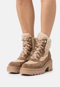 Bottes de randonnée en suède marron et cuir beige avec un col en polaire, une semelle épaisse et colorée et des lacets multicolores. Accents en matériel doré.