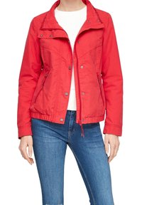 Femme portant une veste zippée rouge sur une chemise blanche avec un jean bleu, debout, les mains détendues le long du corps, sur un fond uni.