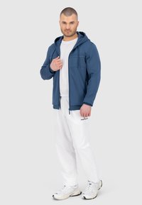 Felpa con zip per uomo in blu navy, caratterizzata da pannelli testurizzati e tasche frontali, abbinata a pantaloni bianchi e scarpe sportive.