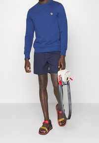 Sudadera azul, pantalones cortos marino, sandalias multicolores con acentos rojos. Sosteniendo una pequeña bolsa blanca con correas rojas y azules.