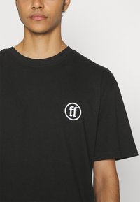 Schwarzes Baumwoll-T-Shirt mit Kurzarm, Rundhalsausschnitt und einem kreisförmigen weißen Logo mit "ff" im linken Brustbereich.