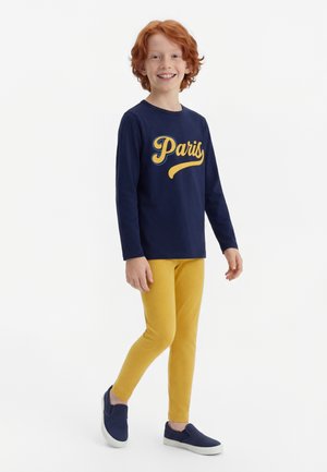 Red-haired child stands smiling, wearing navy blue long-sleeve shirt with "Paris" text, mustard yellow pants, and navy slip-on shoes.