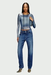 Top de manga comprida metálico azul, combinado com jeans azuis de cintura alta e corte largo, e sapatos verdes de bico fino. Pequena mala segurada com uma mão.