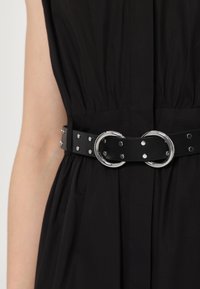 Cintura in pelle nera con due fibbie circolari in argento e borchie decorative in argento, indossata sopra un indumento scuro con dettagli di tessuto arricciato.