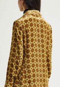 Camicia a maniche lunghe con un motivo decorativo in giallo e marrone, con forme geometriche e dettagli floreali. Tessuto liscio con collo a camicia.